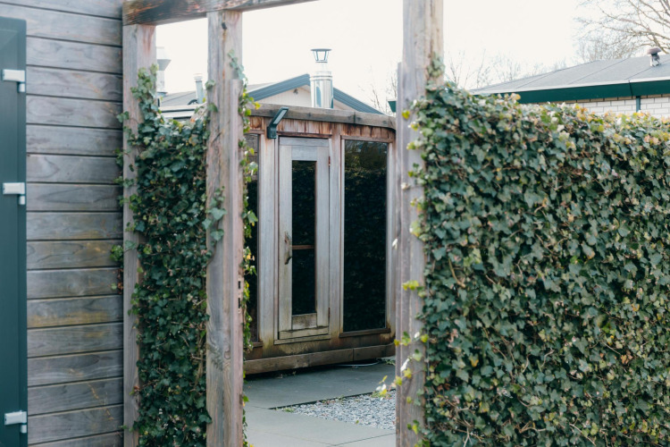 Entrada a la sauna exterior de Veluwe Villa en el parque vacacional De Boshoek, Países Bajos, con setos verdes.