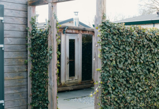 Entrada a la sauna exterior de Veluwe Villa en el parque vacacional De Boshoek, Países Bajos, con setos verdes.