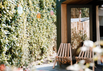 Area esterna della Tiny Wellness Cabin al Holiday Park De IJsvogel con sedie in legno e siepe verde.