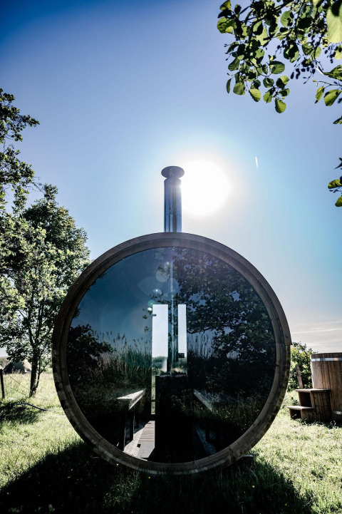 Sauna rotonda in legno con vetro riflettente, sole e alberi, presso TinyParks Cast Away, Paesi Bassi.