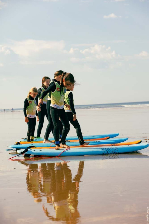 Børn lærer at surfe på stranden nær Les Sables-d'Olonne, iført dragter og stående på surfbrætter.