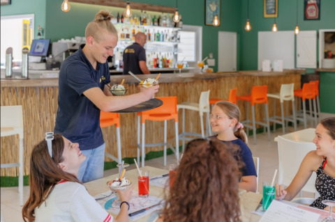 Un camarero sirve helado a clientes sonrientes en una moderna cafetería del Camping Le Bois de la Gachère, Francia.