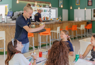 Ein Kellner serviert Eis an fröhliche Gäste in einem modernen Café im Camping Le Bois de la Gachère, Frankreich.