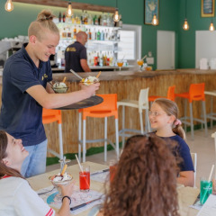 En tjener serverer is til smilende gæster i en moderne café på Camping Le Bois de la Gachère i Frankrig.