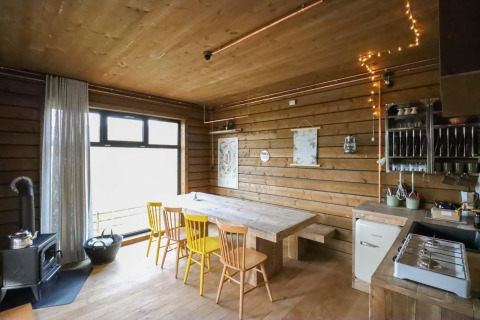 Cocina y comedor con paneles de madera, sillas amarillas, ventana grande y luces acogedoras en la cabaña.