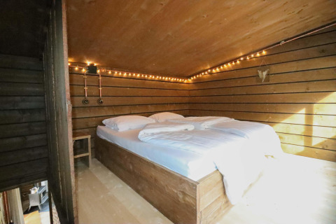 Dormitorio acogedor de tiny house con cama doble, paredes de madera y luces cálidas en el bosque de Bélgica.