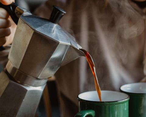 Een persoon schenkt hete koffie uit een mokapot in een groene mok en creëert zo een gezellige sfeer voor glamping ontspanning.