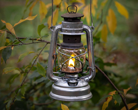 Une lanterne à pétrole vintage avec une flamme allumée suspendue à une branche d'arbre, entourée de feuilles vertes et jaunes.
