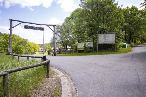 Entrance to Warredal glamping site, featuring a wooden sign, lush greenery, and a paved road under blue skies.
