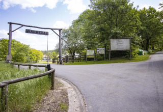 Ingang van het Warredal glampingdomein, met een houten bord, weelderig groen en een verharde weg onder een blauwe lucht.