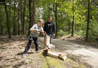 En dreng hugger træ med en økse på en træstub i en skov under opsyn af en voksen på en glampingtur.