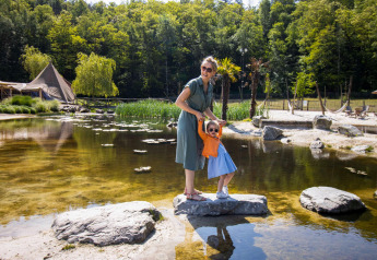 Una madre y su hijo con gafas de sol sobre unas rocas junto a un pintoresco camping con tiendas y un estanque.