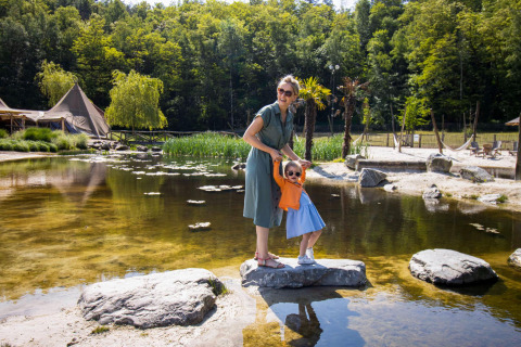 Mutter und Kind mit Sonnenbrille stehen auf Felsen neben einem malerischen Glampingplatz mit Zelten und Teich.