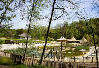 Una vista panorámica de un glamping con tiendas, cabañas de madera y un estanque rodeado de árboles y vegetación.