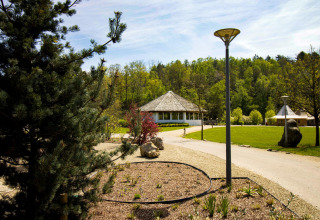 A tranquil glamping site with a round cabin, lush greenery, walking paths, and street lamps under a sunny sky.