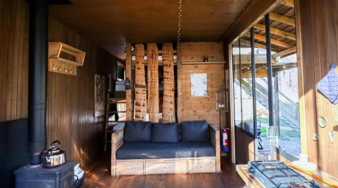 Interior de una cabaña acogedora con paredes de madera, sofá, estufa de leña y grandes ventanales.