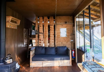 Innenraum einer gemütlichen Lodge mit Holzwänden, Sofa, Holzofen und großen Fenstern zur Natur.