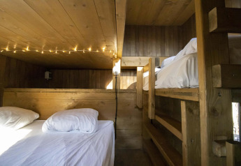 Alloggio glamping accogliente con letti a castello in legno, lenzuola bianche e luci decorative soffuse.