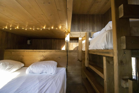 Knusse glampingaccommodatie met houten stapelbedden, wit beddengoed en sfeerverlichting aan het plafond.