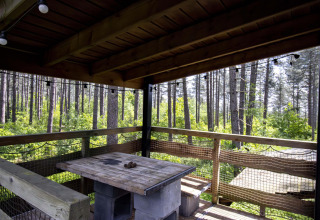 Overdekt terras van een lodge met houten tafel, bank en net, met uitzicht op een uitgestrekt dennenbos.