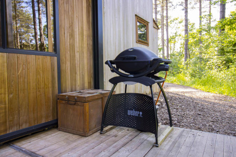 Una barbacoa Weber negra sobre una terraza de madera junto a una cabaña rodeada de bosque verde.