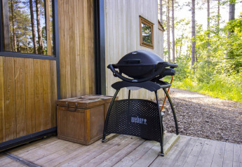 Un barbecue Weber nero si trova su un ponte di legno vicino a una cabina immersa nel verde bosco.