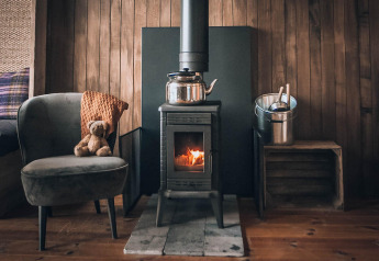 Cozy glamping setup with wood stove, armchair and teddy bear, and metal bucket in a wooden cabin.