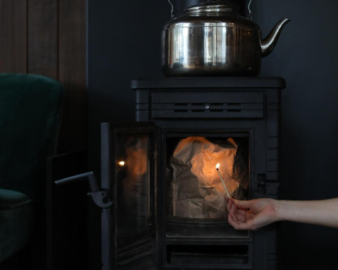 A hand lights a match inside a stove with crumpled paper and a metal kettle on top at glamping lodgings.