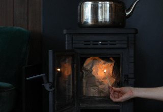 A hand lights a match inside a stove with crumpled paper and a metal kettle on top at glamping lodgings.