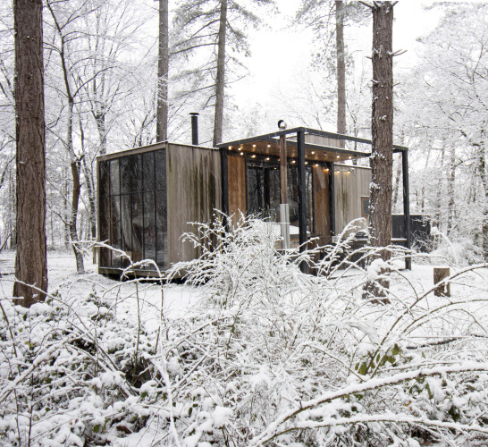 Cabaña cubierta de nieve con grandes ventanales y luces, situada en el bosque de Limburg, Bélgica.