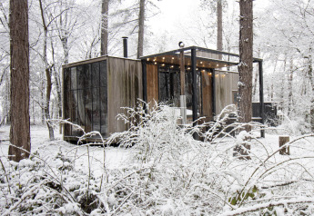 Sneeuwbedekte cabine met grote ramen en sfeerverlichting in het bos van Limburg, België.