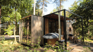 Cabaña moderna de madera en el bosque con ventanas grandes, jacuzzi y zona de comedor en la terraza.
