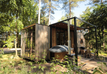 Cabina moderna in legno nella foresta con ampie vetrate, vasca idromassaggio e zona relax esterna.