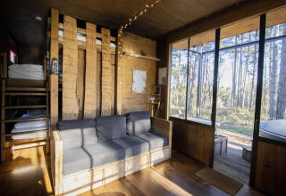 Interior de una cabaña acogedora con literas, sofá gris y grandes ventanales que dan al bosque en Limburgo.