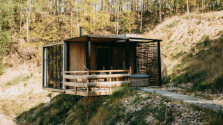 Cabina moderna junto al agua con bañera de hidromasaje en la terraza, rodeada de bosque y colinas.