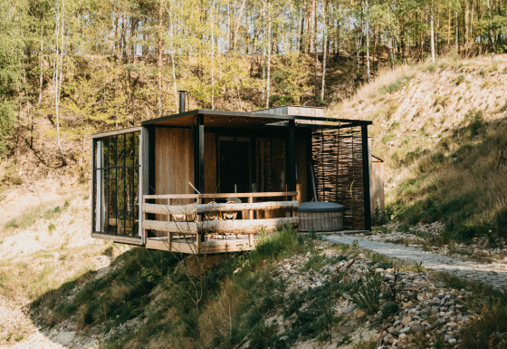 Modern water cabin with hot tub on the deck, surrounded by natural landscape and forest hills.