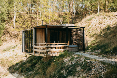 Modern water cabin with hot tub on the deck, surrounded by natural landscape and forest hills.