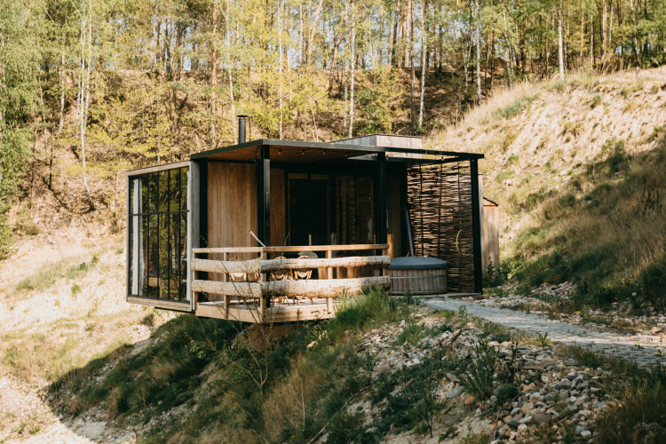 Cabina moderna junto al agua con bañera de hidromasaje en la terraza, rodeada de bosque y colinas.