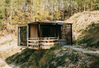 Cabina moderna junto al agua con bañera de hidromasaje en la terraza, rodeada de bosque y colinas.