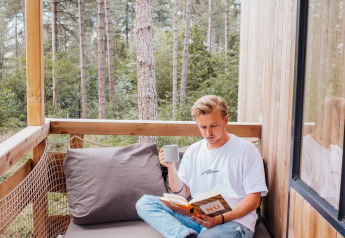 Mand læser en bog og drikker kaffe på en hyggelig balkon i et træhus i Limburgs skov, Belgien.