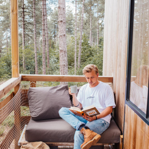 Mand læser en bog og drikker kaffe på en hyggelig balkon i et træhus i Limburgs skov, Belgien.