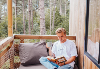 Man leest een boek en drinkt koffie op het balkon van een boomhut in het bos van Limburg, België.