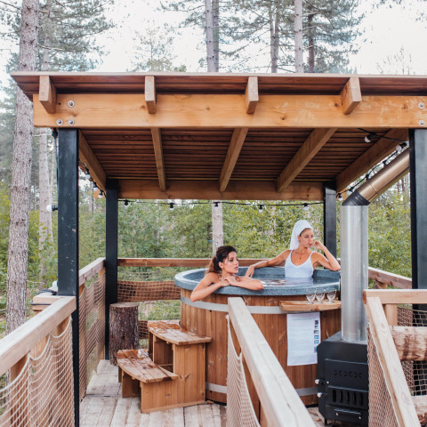 Twee vrouwen ontspannen in een buiten bubbelbad op een boomhut terras in het bos van Limburg, België.