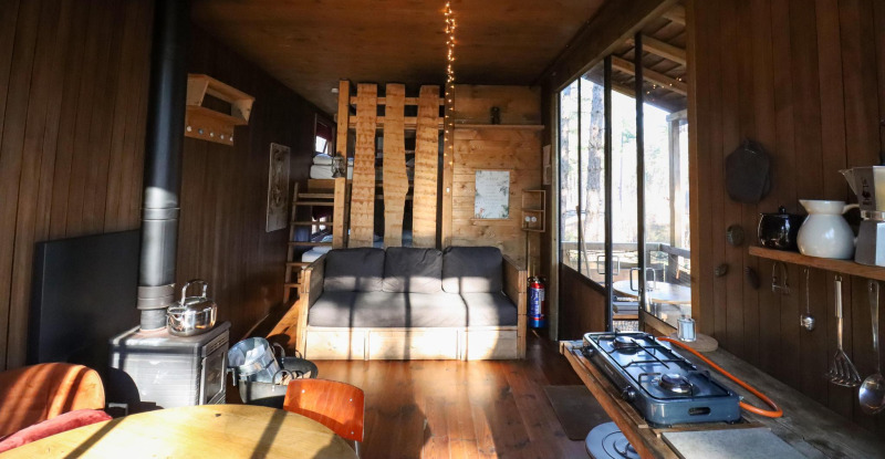 Acogedora casa en el árbol con sofá, estufa de leña, cocina pequeña y grandes ventanas al bosque.