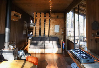 Gemütliches Baumhaus mit Sofa, Holzofen, kleiner Küche und großen Fenstern mit Blick ins Grüne.