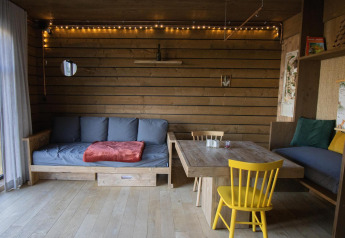 Knusse cabin in Limburg, België, met houten interieur, sofa, eettafel en gele stoelen in het bos.
