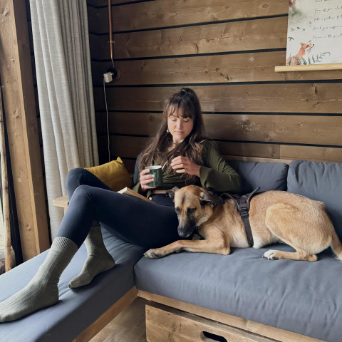Mujer relajándose con una taza de té y un perro en un sofá gris dentro de una cabaña de madera en Limburg, Bélgica.