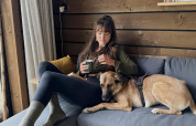Mujer relajándose con una taza de té y un perro en un sofá gris dentro de una cabaña de madera en Limburg, Bélgica.