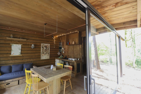 Acogedora cabaña con interior de madera, mesa de comedor y amplias ventanas con vistas al bosque.