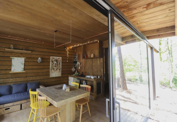 Interno accogliente di una cabina in legno con tavolo da pranzo, divano e grandi finestre sul bosco.